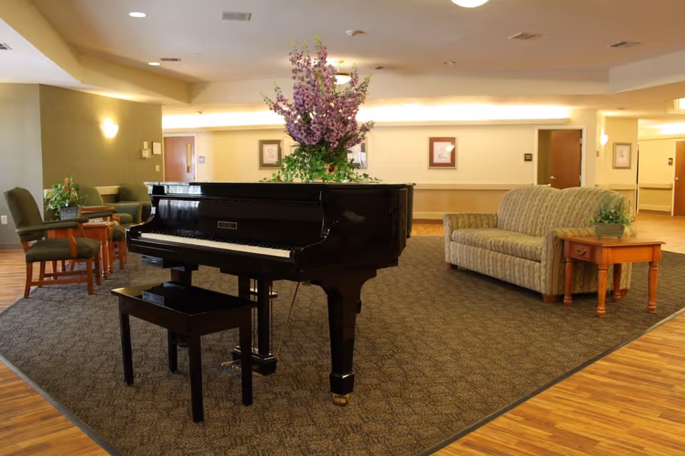 A spacious and well-lit common area featuring a black grand piano with a floral arrangement on top, a patterned sofa, wooden side tables with potted plants, and several chairs arranged around the room. The floor is a combination of carpet and wood, and the walls are decorated with framed pictures and soft lighting.