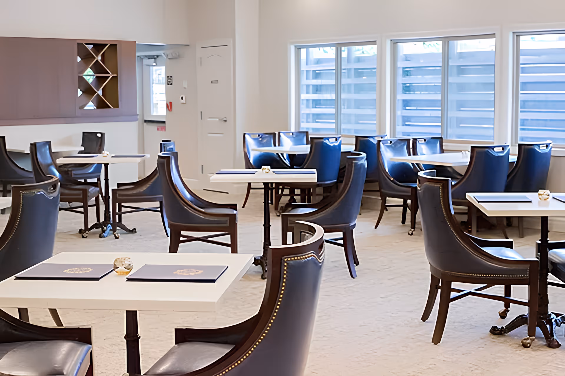 A bright dining room with multiple white tables and dark leather chairs arranged neatly. The room has large windows letting in natural light and a clean, modern design with light-colored walls and carpeted floor.