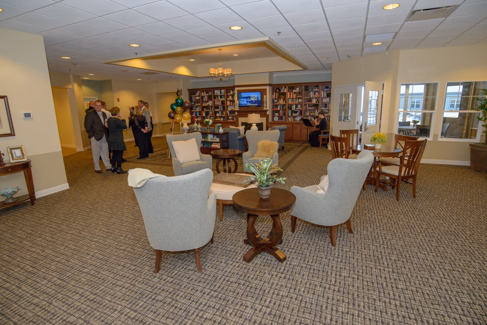 A spacious senior living facility common area with several comfortable armchairs arranged around small wooden tables. In the background, a group of people are conversing near a bookshelf filled with books and a television mounted above a fireplace. There is a person playing a piano near the bookshelf. The room has large windows letting in natural light and a carpeted floor.