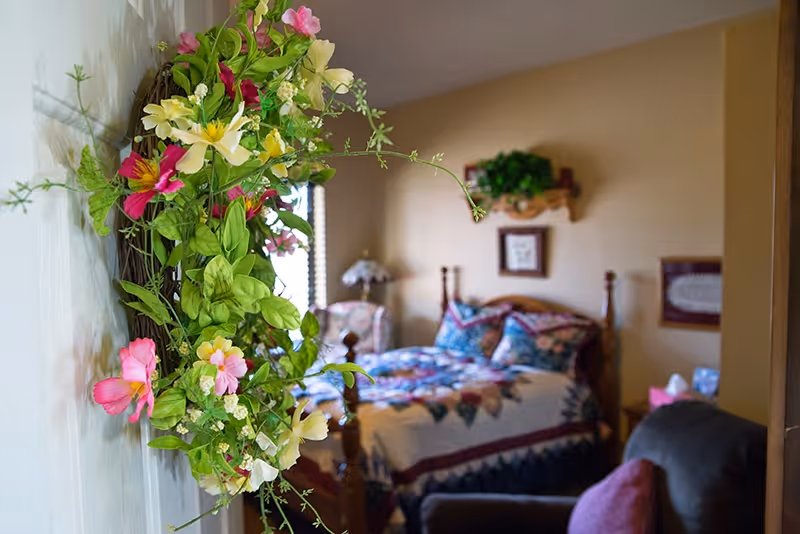 A cozy bedroom with a wooden bed covered in a colorful quilt and pillows. There is a floral wreath hanging on the wall in the foreground, and a comfortable armchair with a pink cushion is visible. The room has warm beige walls with framed pictures and a small shelf with a plant.