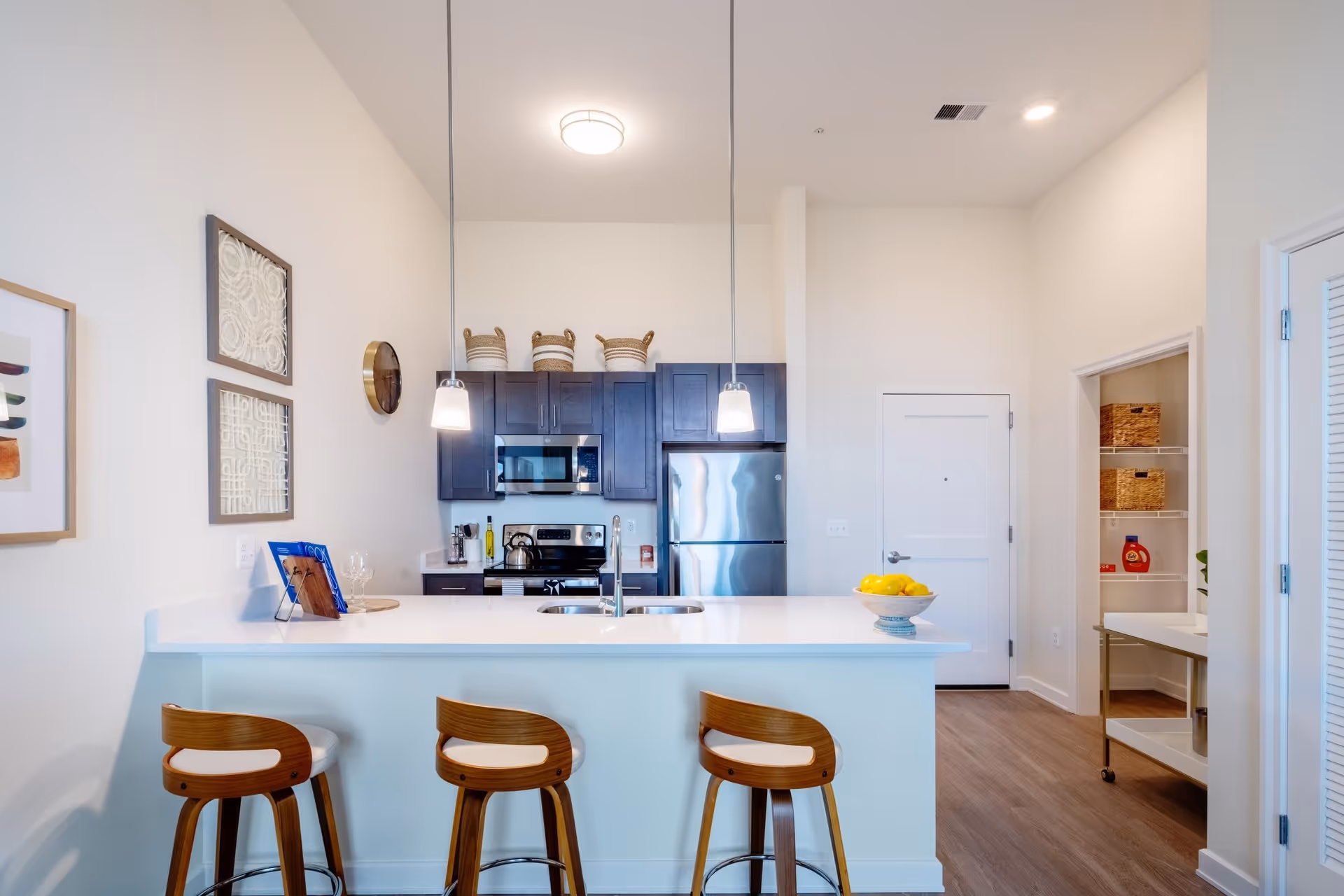 Modern kitchen with a white island countertop and three wooden bar stools. Above the island, two pendant lights hang from the ceiling. The kitchen features dark cabinets, a stainless steel refrigerator, microwave, and stove. On the right side, there is a pantry with shelves holding baskets and a bottle of detergent. The walls are white, and the floor has a wood finish.