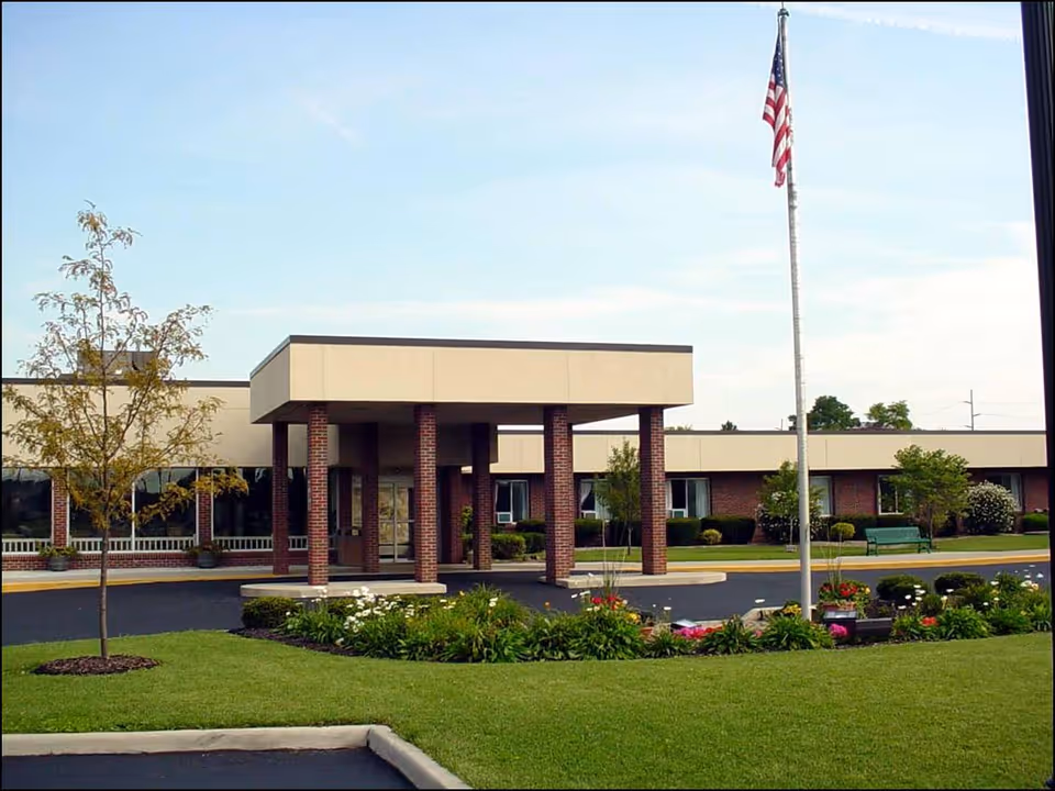 Exterior view of Wood Haven Healthcare building with a covered entrance supported by brick pillars, a flagpole with an American flag, landscaped garden beds with flowers and shrubs, and a well-maintained lawn.