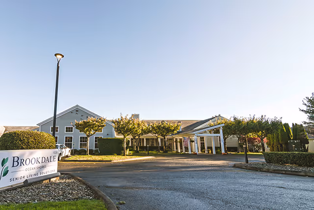 Exterior view of a senior living facility building with a driveway, small trees, and a sign that reads 'Brookdale Ocean Shores Senior Living Solutions' under a clear blue sky.