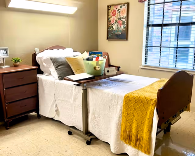 A neatly made hospital-style bed with white bedding and a yellow throw blanket in a well-lit room. The bed has a wooden headboard and footboard, with a small overbed table holding a green basket filled with books and other items. Next to the bed is a wooden nightstand with a small plant and a framed photo. A window with blinds and a floral painting on the wall are visible in the background.