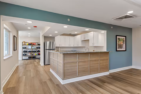 Modern kitchen area with a large island featuring a granite countertop and wood paneling. The kitchen has white cabinets, a stainless steel refrigerator, and a sink. The walls are painted in a combination of white and teal, with recessed lighting in the ceiling. In the background, there is a shelving unit with various items and a window letting in natural light.