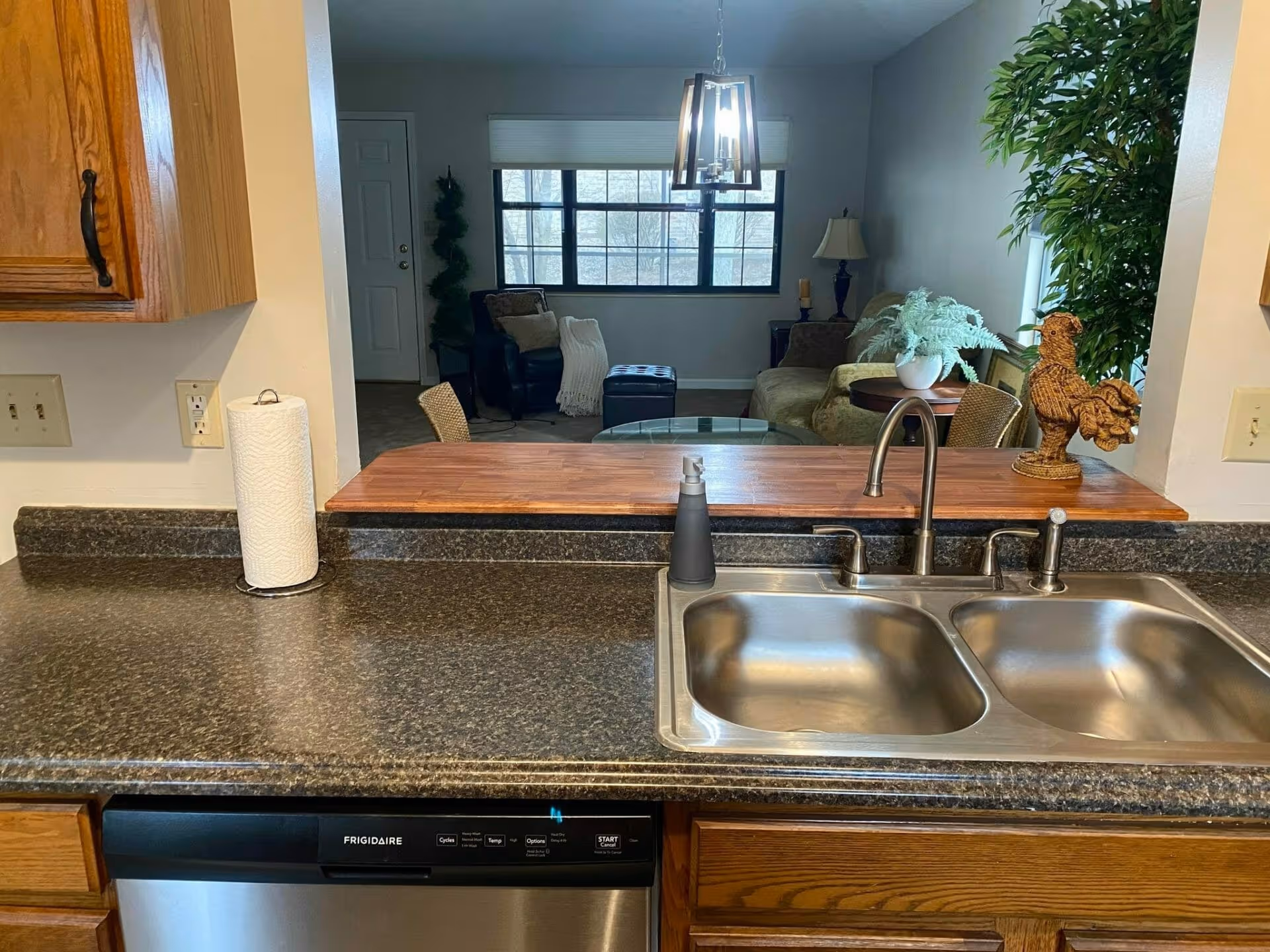 View of a kitchen sink with a double basin stainless steel sink, a soap dispenser, and a paper towel holder on a dark granite countertop. Behind the sink is a wooden counter ledge with two chairs, a decorative rooster figurine, and a potted plant. In the background, there is a living room area with a sofa, armchair, side table with a lamp, and a large window letting in natural light.