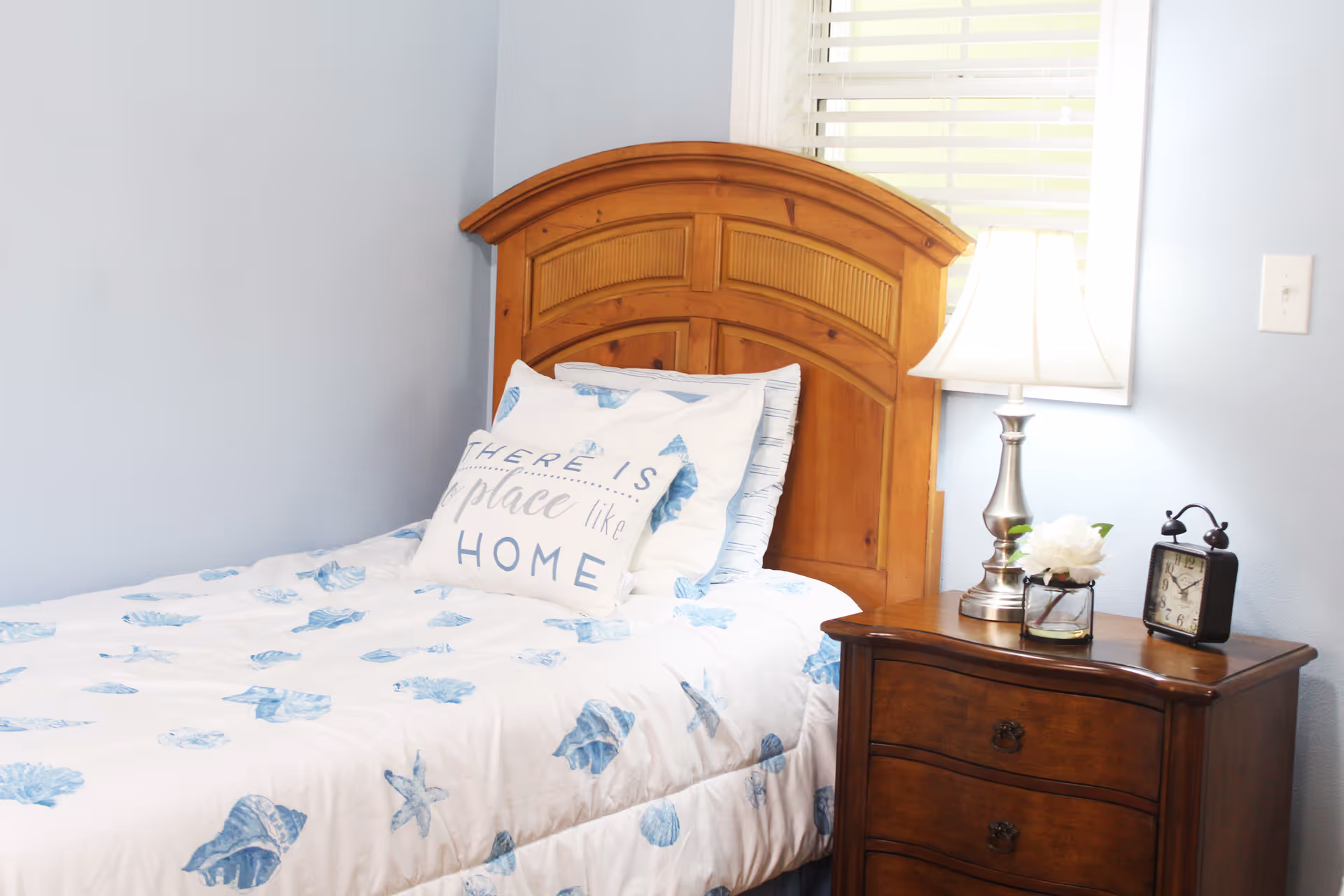 Single bed with a wooden headboard, blue shell-patterned bedding and a pillow reading 'HOME', next to a nightstand with a lamp and alarm clock.