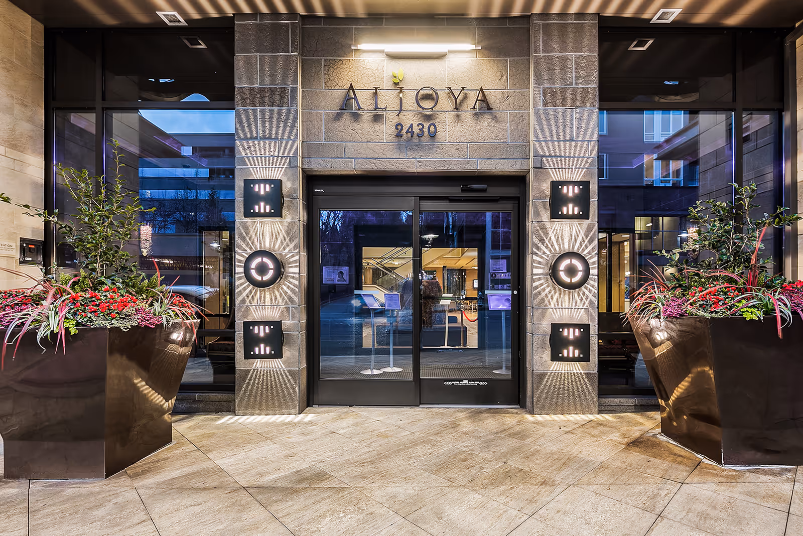 Entrance to Aljoya Mercer Island featuring an illuminated "ALJOYA 2430" sign above glass automatic doors flanked by large planters and decorative lighting.