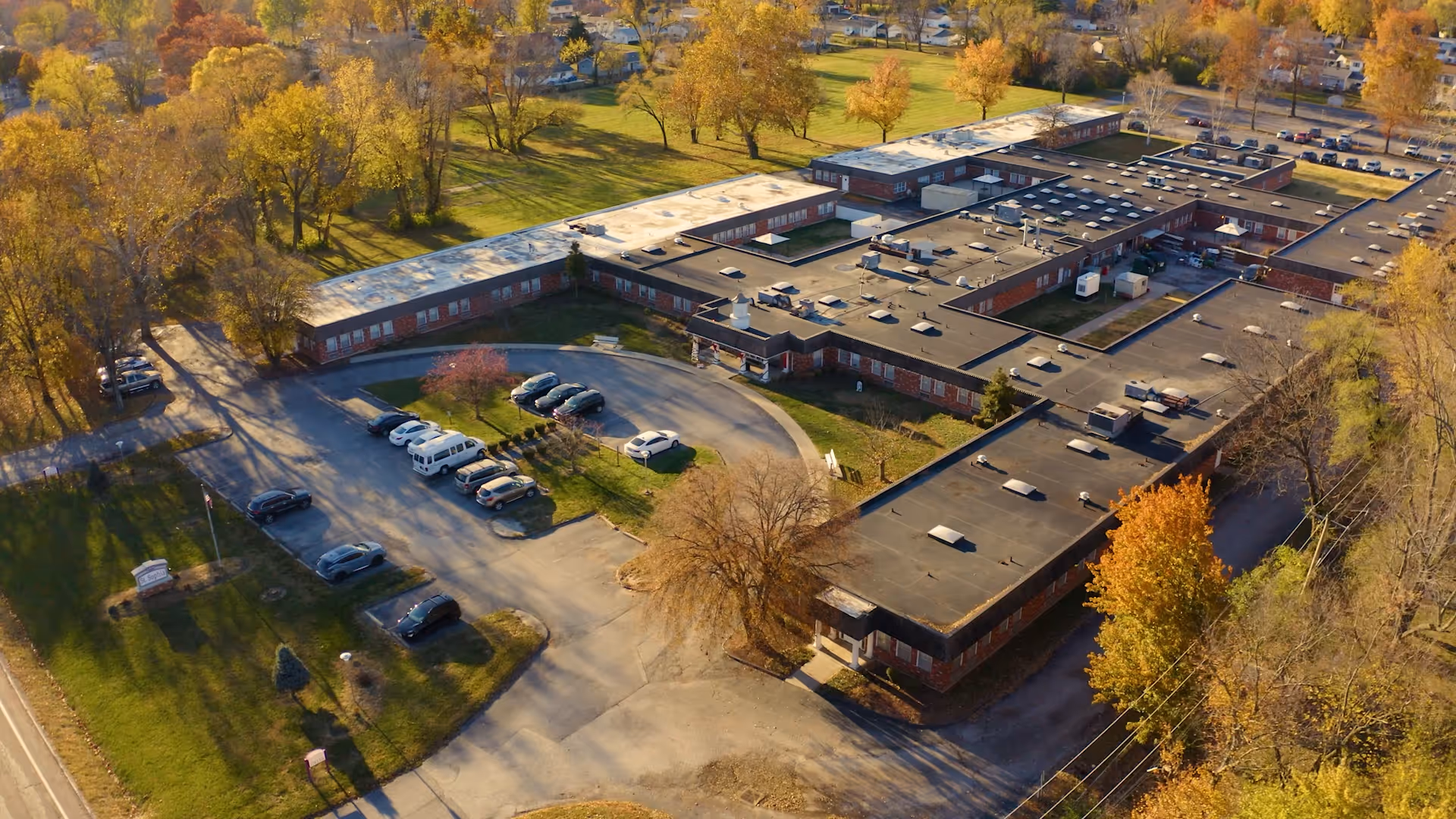 Aerial view of St Sophia Health & Rehabilitation Center, a large single-story building with a flat roof surrounded by trees with autumn foliage. Several cars are parked in the parking lot and driveway areas around the facility. The surrounding area includes grassy lawns and residential neighborhoods.