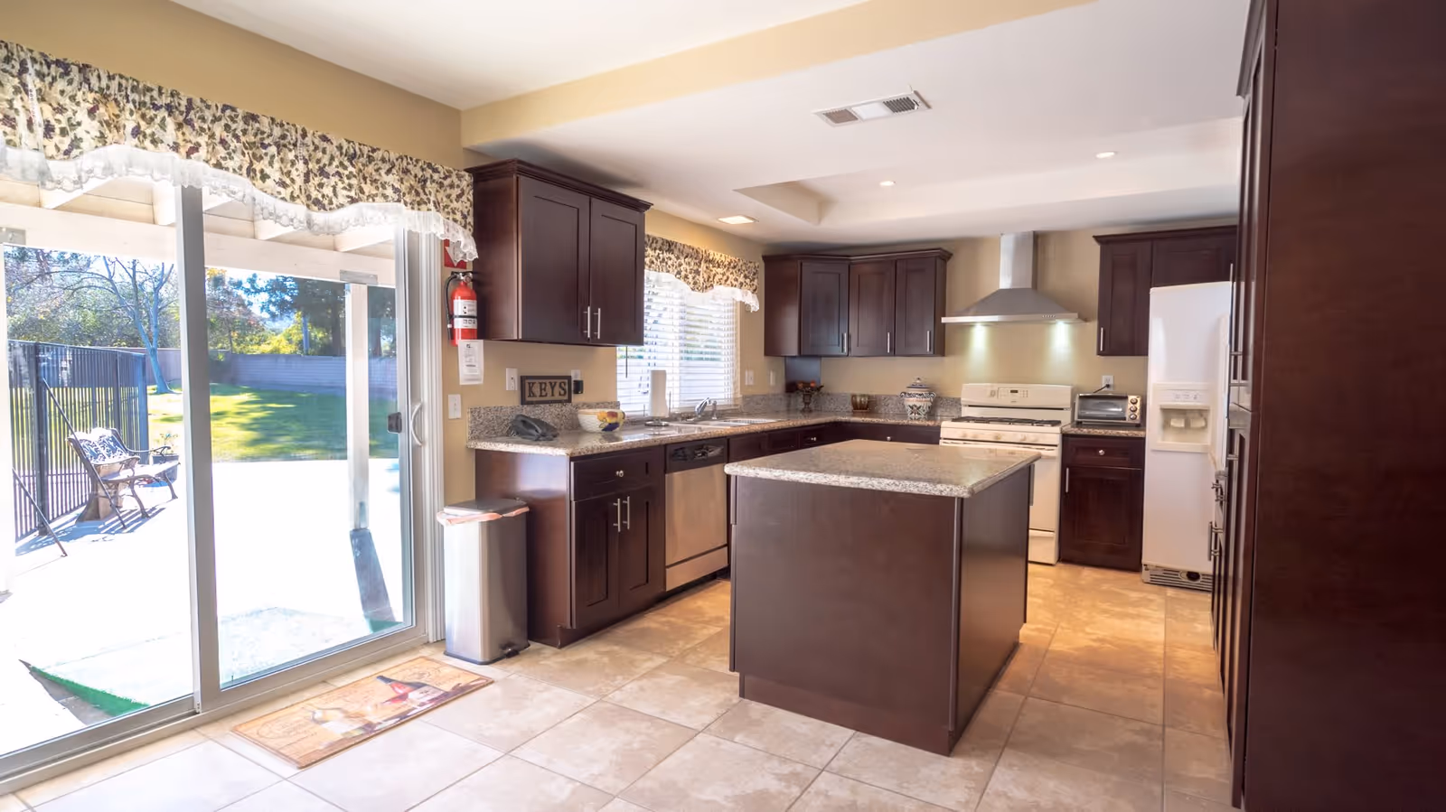 Bright kitchen with dark wooden cabinets, granite countertops, a central island, white stove with a stainless steel hood, dishwasher, and refrigerator. There is a sliding glass door leading to a green outdoor area with a bench and trees. Floral valances adorn the windows.