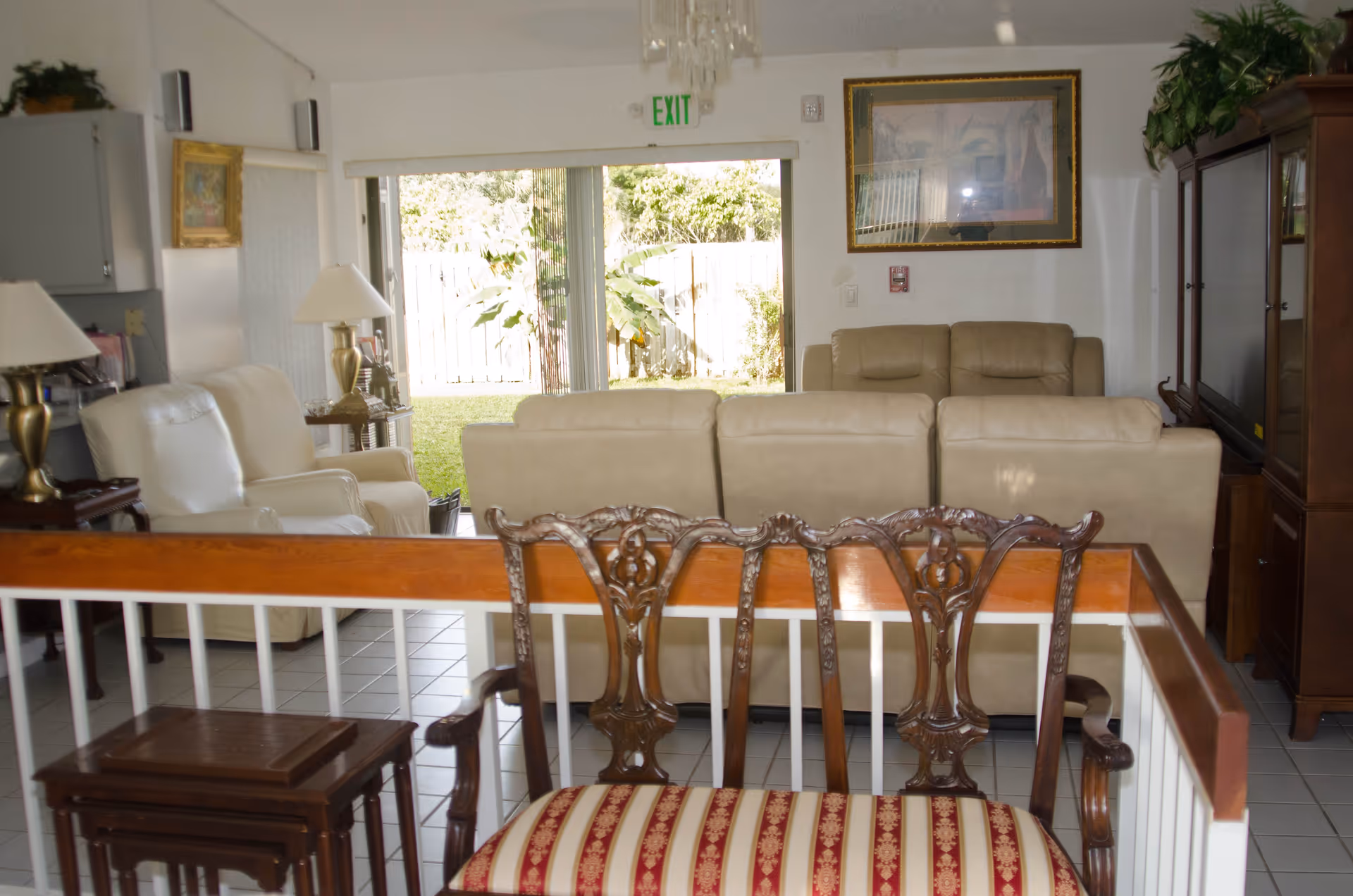 Interior view of a living room area in a senior living facility with beige leather sofas, a wooden chair with red and gold upholstery in the foreground, a large TV cabinet on the right, two table lamps on side tables, framed artwork on the walls, and a sliding glass door leading to a green outdoor space.