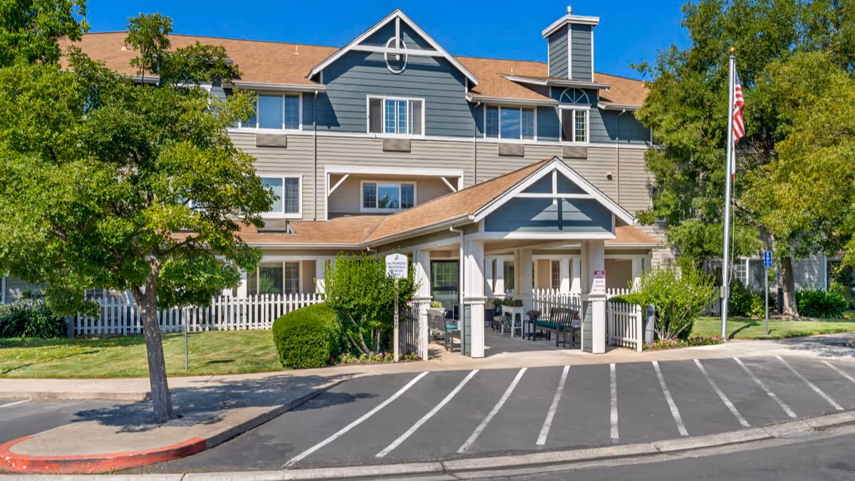 Exterior view of a senior living facility building with a covered entrance, surrounded by trees and greenery, with a parking area in front and an American flag on a flagpole to the right.