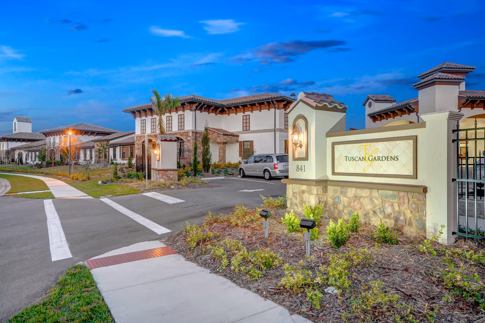 Exterior view of Tuscan Gardens Of Venetia Bay facility at dusk, showing a well-lit entrance sign with the facility name, a driveway, landscaped garden beds, and a large building with Mediterranean-style architecture featuring tiled roofs and stone accents.
