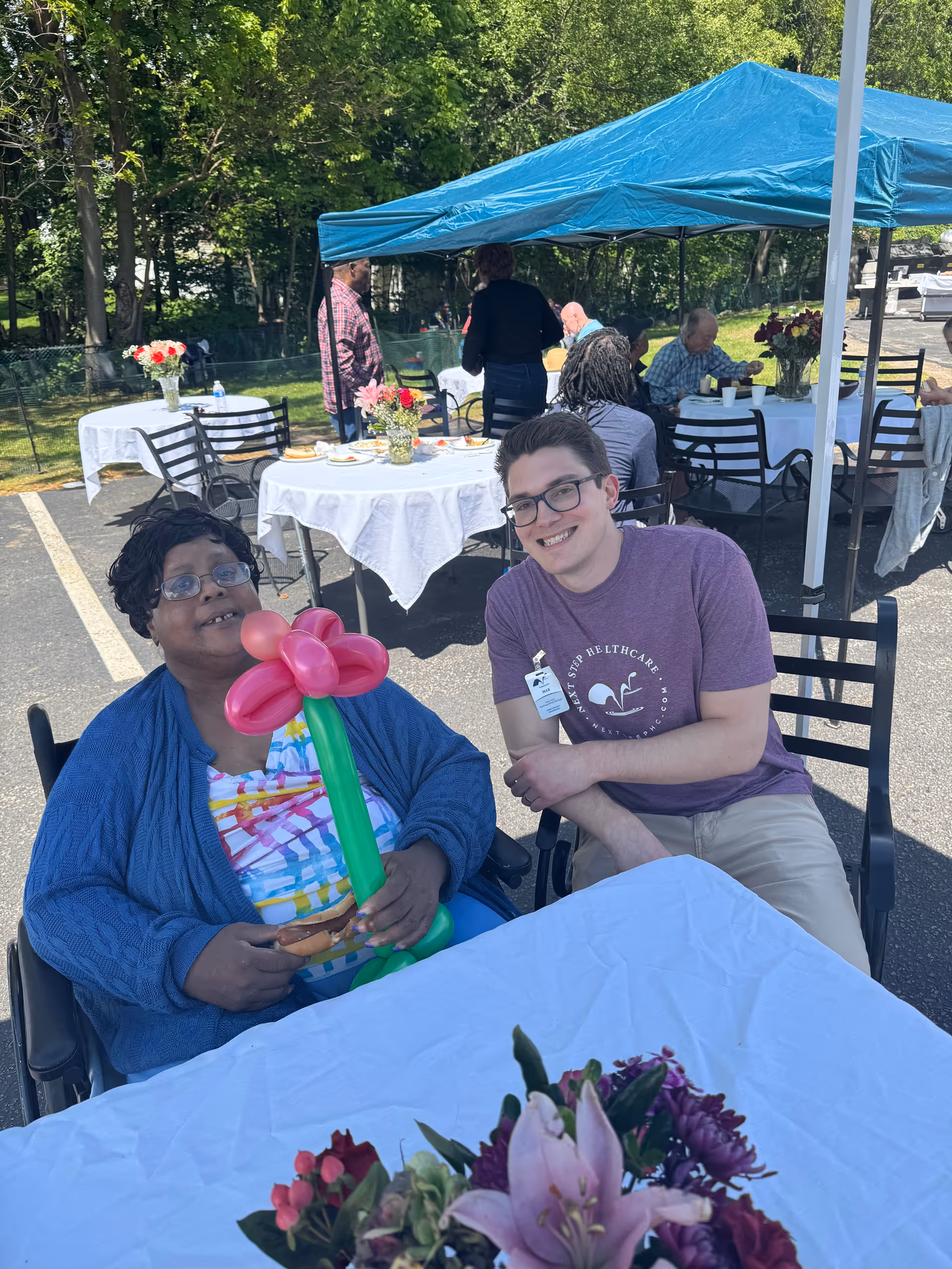 An older woman holding a balloon flower sits next to a smiling young man at an outdoor table during a canopy-covered gathering.