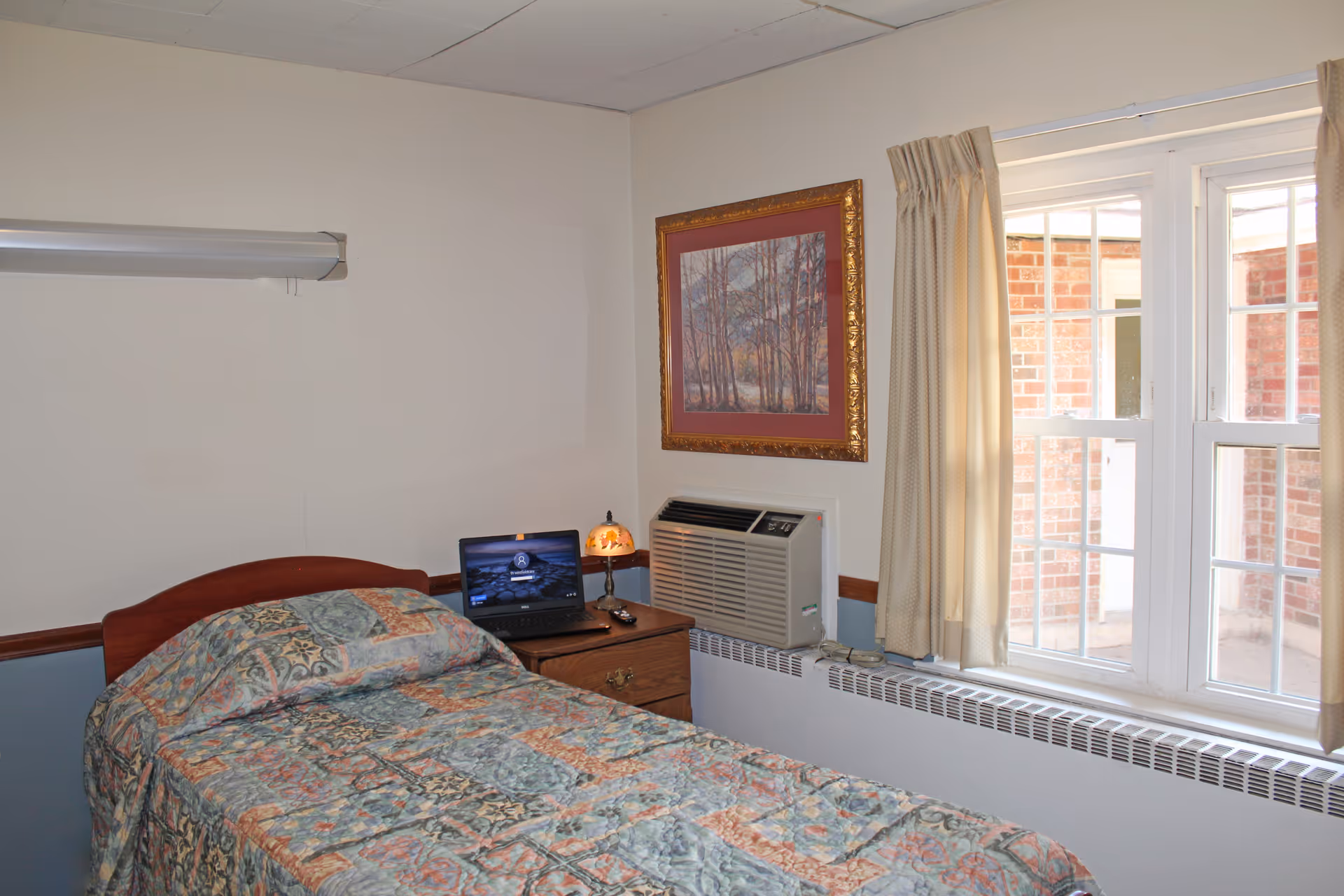 A small bedroom with a single bed covered in a patterned quilt. Next to the bed is a wooden nightstand with a laptop, a small lamp, and a remote control. There is a window with beige curtains letting in natural light, and an air conditioning unit installed below the window. A framed picture of trees hangs on the wall above the air conditioner.