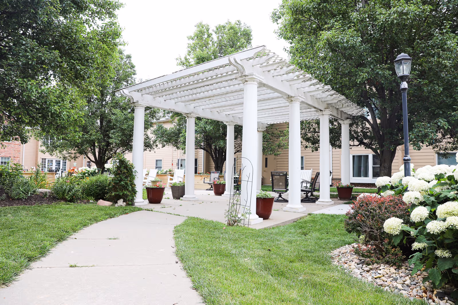 Outdoor garden area with a white pergola supported by columns, surrounded by green grass, trees, flowering plants, and a paved walkway leading to the pergola. There are chairs and potted plants under the pergola, and a beige building with windows in the background.