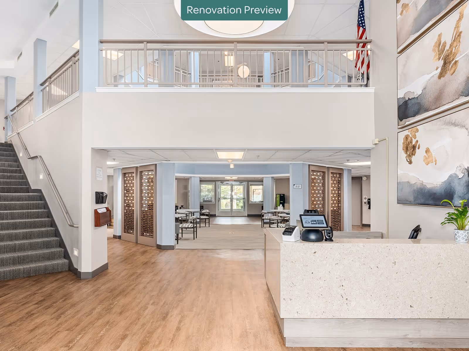 Interior view of a senior living facility lobby area with a reception desk on the right, a staircase with carpet on the left, and a dining area with tables and chairs visible through an open space in the center. The space features light wood flooring, neutral walls, and large abstract paintings on the right wall. An American flag is displayed near the upper railing, and a sign above reads 'Renovation Preview'.