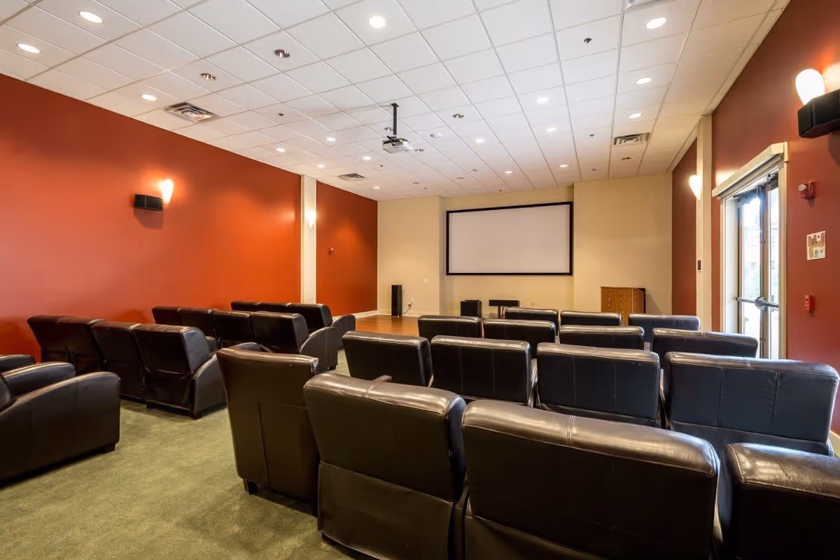 A small theater room with several rows of black leather recliner chairs facing a large projection screen on a beige wall. The room has red and beige walls, green carpet, ceiling lights, and a projector mounted on the ceiling. There is a door with glass panels on the right side of the room.