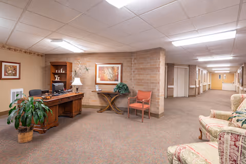 Reception-style seating area in a nursing facility hallway with a wooden desk, chairs, artwork, and plants.