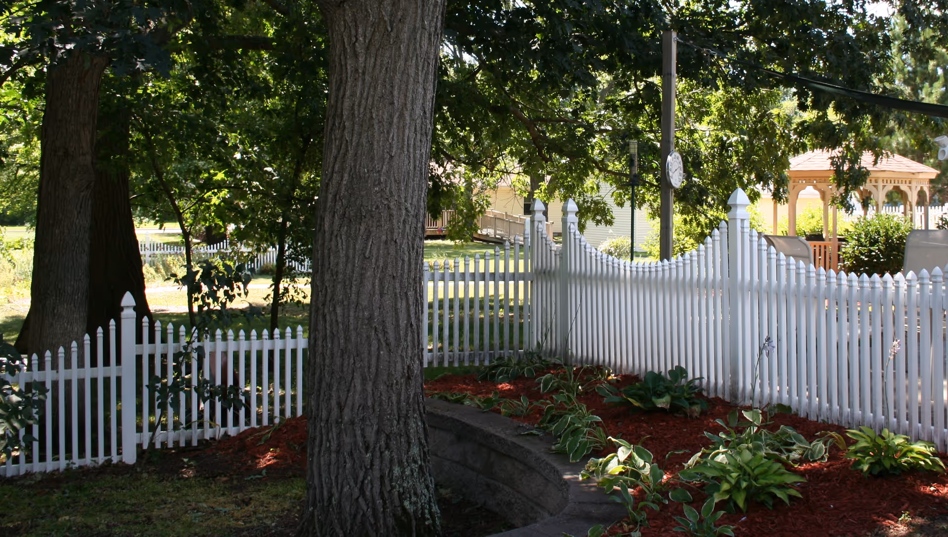 A garden area with a white picket fence, large trees providing shade, and a gazebo in the background. The garden has mulch and green plants along the fence line.
