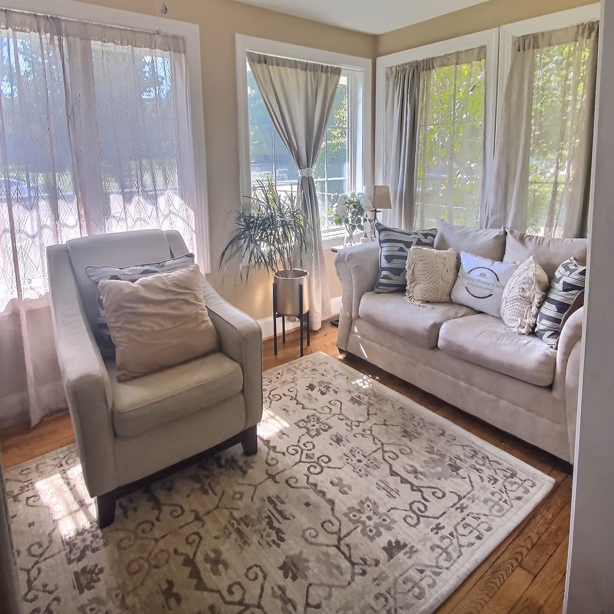A cozy living room with a beige armchair and a matching sofa adorned with multiple decorative pillows. The room has large windows with sheer and solid curtains allowing natural light to fill the space. A patterned area rug covers the wooden floor, and a potted plant and a small lamp sit near the windows.