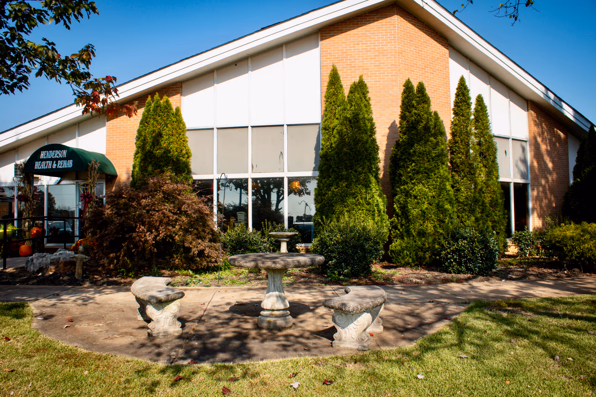 Front exterior of Henderson Health & Rehab building showing a brick facade, tall shrubs, a green entrance awning, and a small stone table with benches in the landscaped courtyard.