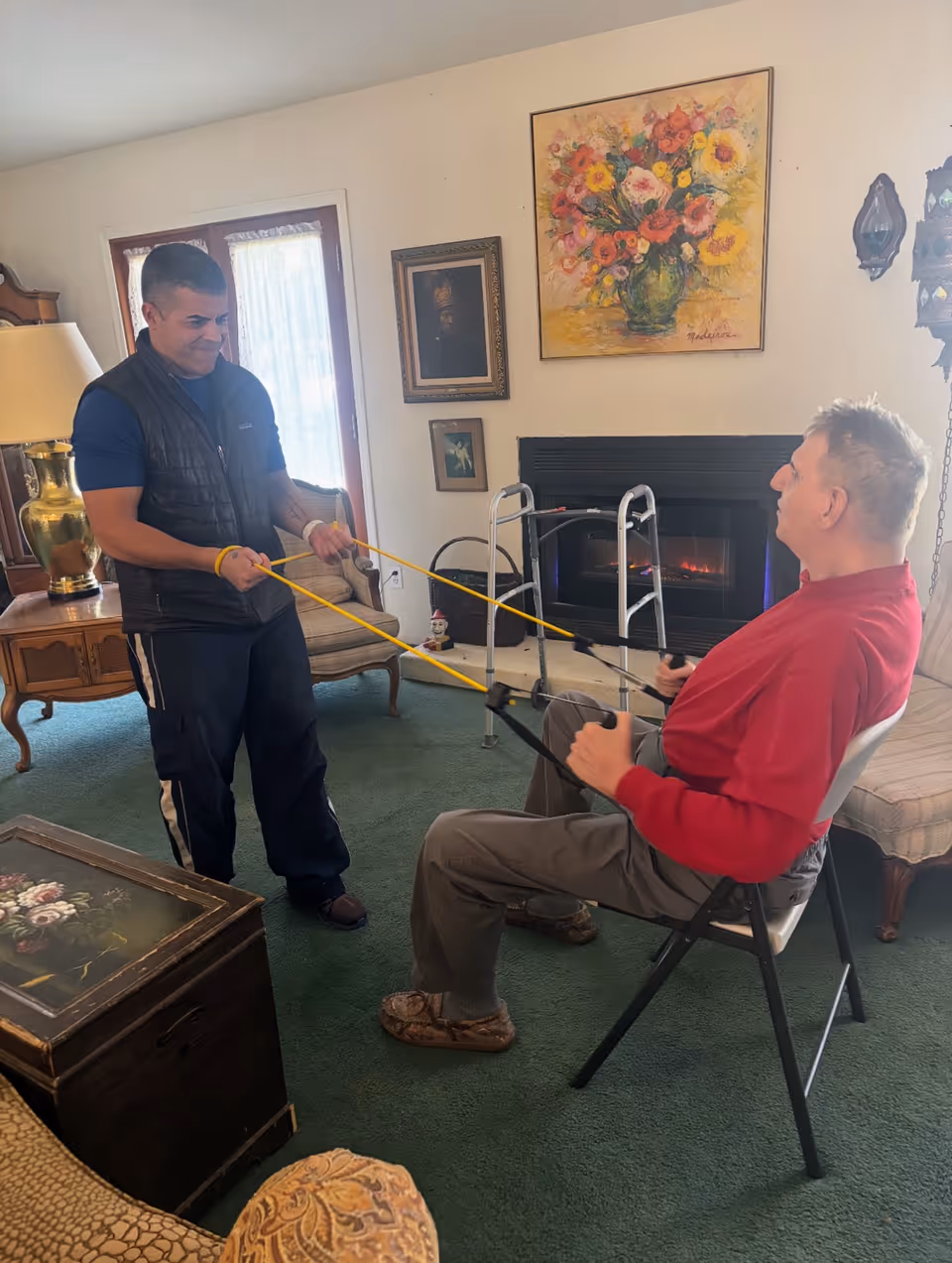 Two men using resistance bands in a cozy living room with a fireplace and floral artwork.