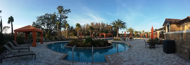 Outdoor swimming pool area with a circular pool surrounded by a paved deck. There are lounge chairs and cabanas with orange curtains on the left side, palm trees and other greenery around the pool, and tables with umbrellas on the right side. The sky is clear with some light clouds.