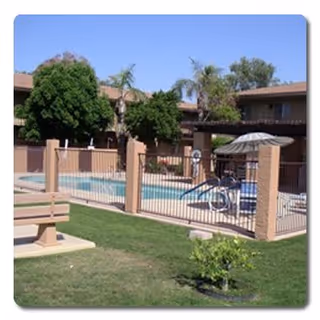 Outdoor area of Desert Marigold Senior Living of Tempe featuring a fenced swimming pool with lounge chairs and an umbrella, surrounded by grass, trees, and a bench in the foreground. Residential buildings are visible in the background under a clear blue sky.