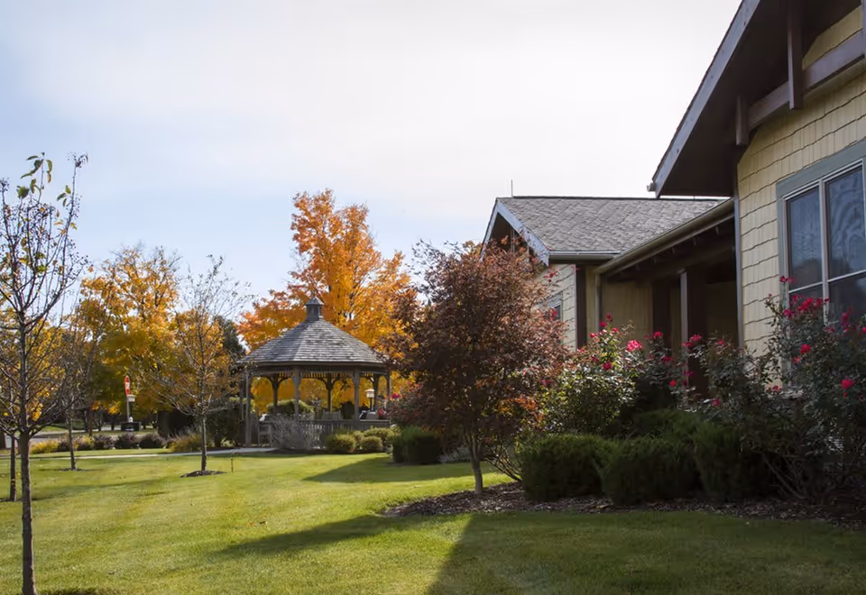 A well-maintained outdoor garden area of a senior living facility with green grass, small trees, bushes with pink flowers, and a wooden gazebo. The building exterior is visible on the right side with beige siding and windows. Trees with autumn-colored leaves are in the background under a clear sky.