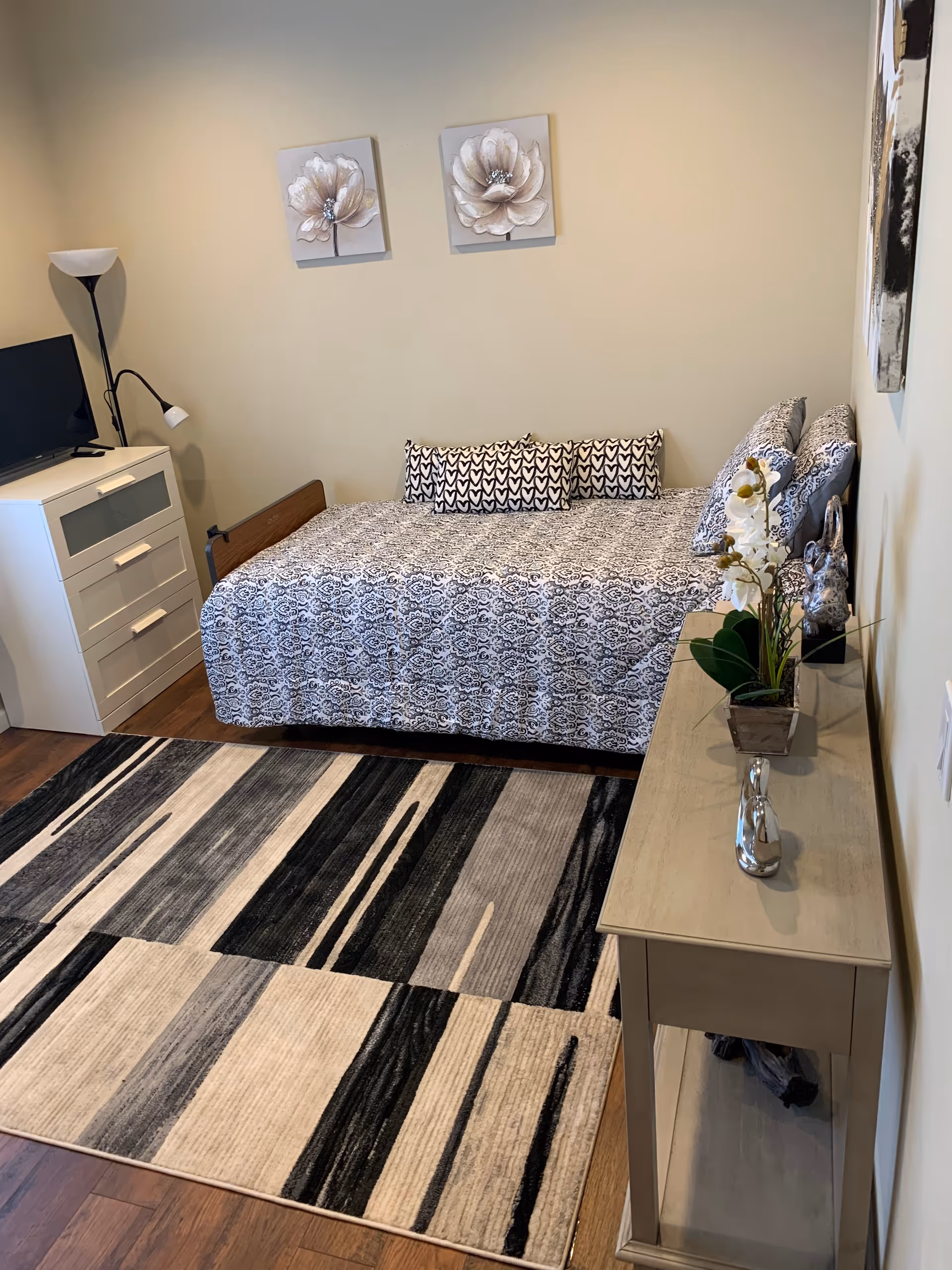 A neatly arranged bedroom with a single bed covered in a patterned black and white bedspread and matching pillows. Above the bed are two floral paintings. To the left of the bed is a white dresser with a small TV and a floor lamp with two lights. On the right side, there is a beige console table with decorative items including a potted orchid and a silver figurine. The floor has a large area rug with a modern black, gray, and beige geometric pattern.