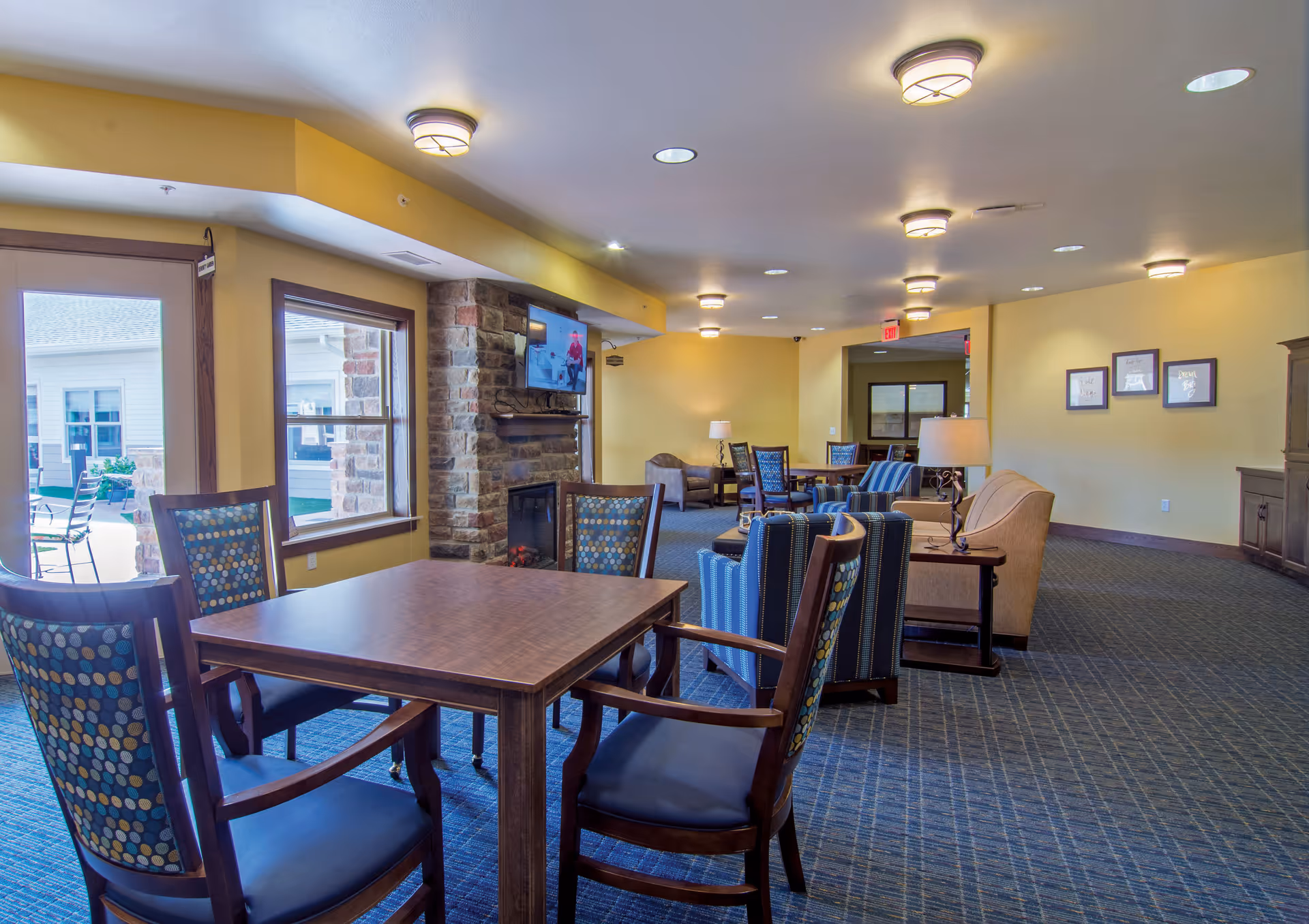 Bright communal lounge and dining area with wooden tables and patterned chairs, a stone fireplace with TV, and additional seating in a senior living facility.