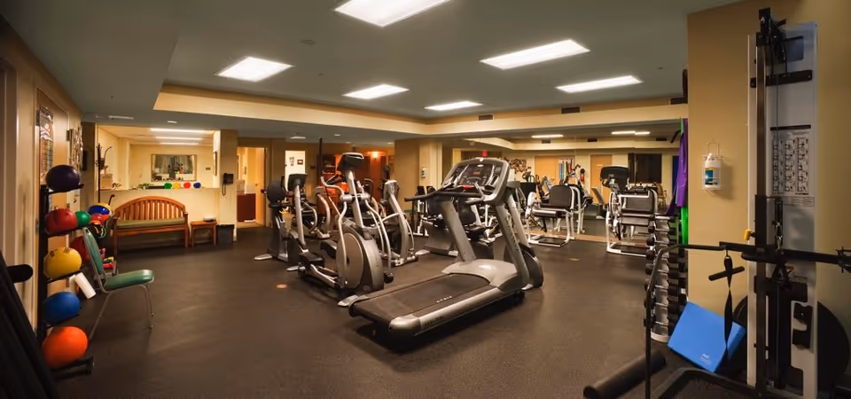 Interior view of a fitness room in a senior living facility featuring various exercise equipment including treadmills, elliptical machines, stationary bikes, and weight machines. The room has a dark floor, beige walls, and bright overhead lighting. There are colorful exercise balls and mats stored on racks along one wall, a wooden bench, and a large mirror covering one side of the room.