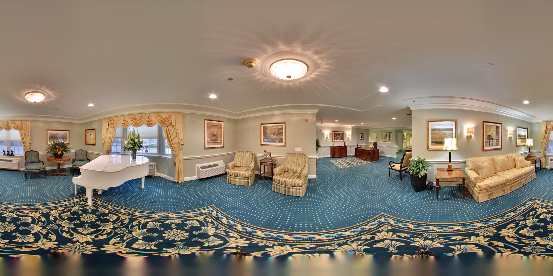 Spacious senior living lounge with a white grand piano, upholstered seating, decorative carpet, and a reception desk in the background.