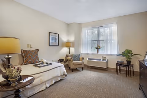 A cozy bedroom in a senior living facility with a single bed adorned with patterned pillows and a tray, a side table with a lamp and decorative items, an armchair with a cushion, a window with sheer curtains letting in natural light, a wall-mounted air conditioning unit below the window, and a small table with a plant and picture frame.