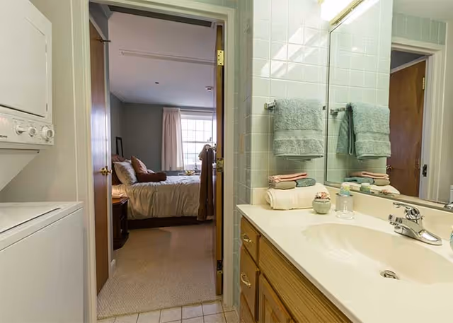 Bathroom vanity with sink and mirror, folded towels, a stacked washer/dryer, and an open door revealing a bedroom.