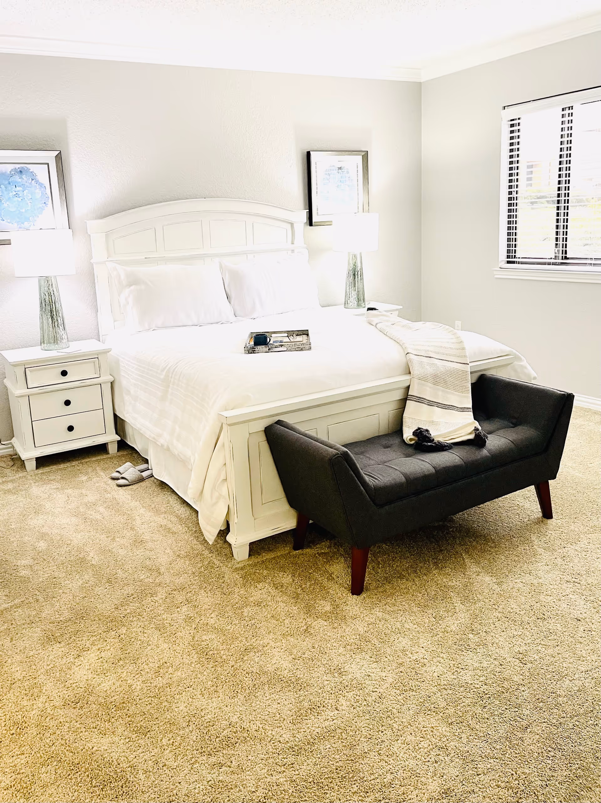 A bright and clean bedroom with a white wooden bed frame and white bedding. There are two bedside tables with matching lamps and framed artwork above them. A dark gray upholstered bench with wooden legs is placed at the foot of the bed, with a folded blanket draped over it. The room has beige carpet flooring and a window with blinds on the right wall.