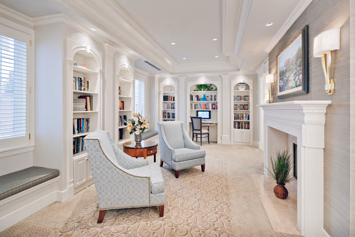 Bright elegant sitting room with built-in bookshelves, two upholstered chairs, a fireplace, and a small desk area.
