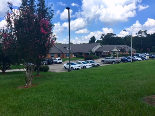Exterior view of Copiah Living Center showing a single-story brick building with a gray roof, a parking lot with several cars, green grass in the foreground, and a tree with pink flowers on the left side under a partly cloudy blue sky.