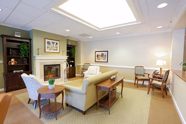 A cozy living room area in a senior living facility with a beige sofa, two patterned armchairs, a wooden side table with a small plant, a fireplace with a framed artwork above it, built-in dark wood shelves with decorative items, and a desk with two chairs and a lamp in the corner.