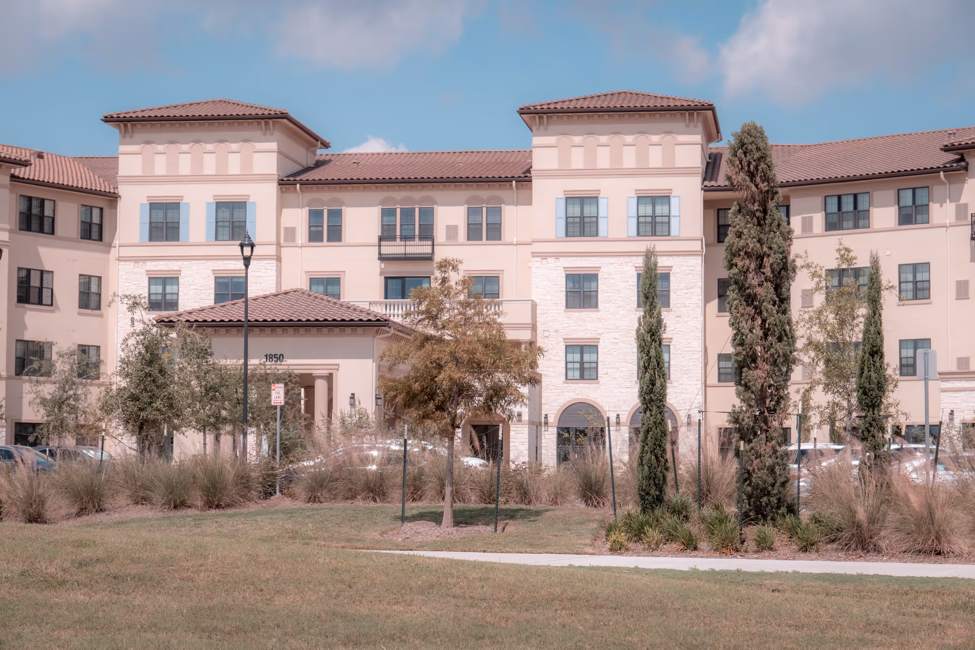 Exterior view of a multi-story senior living facility building with beige walls, stone accents, and a tiled roof. The building is surrounded by landscaped greenery including trees, shrubs, and grass under a partly cloudy blue sky.