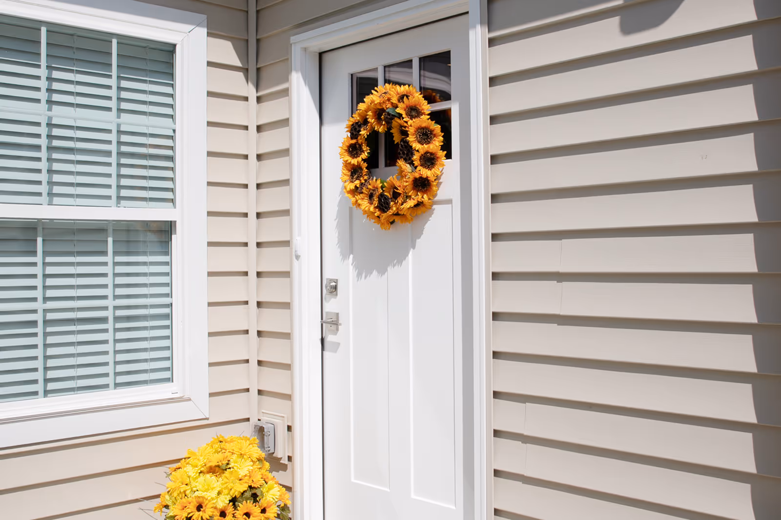 White exterior door with a sunflower wreath hanging on it, next to a window with white blinds and beige siding on the building. A pot of yellow flowers is placed near the door.