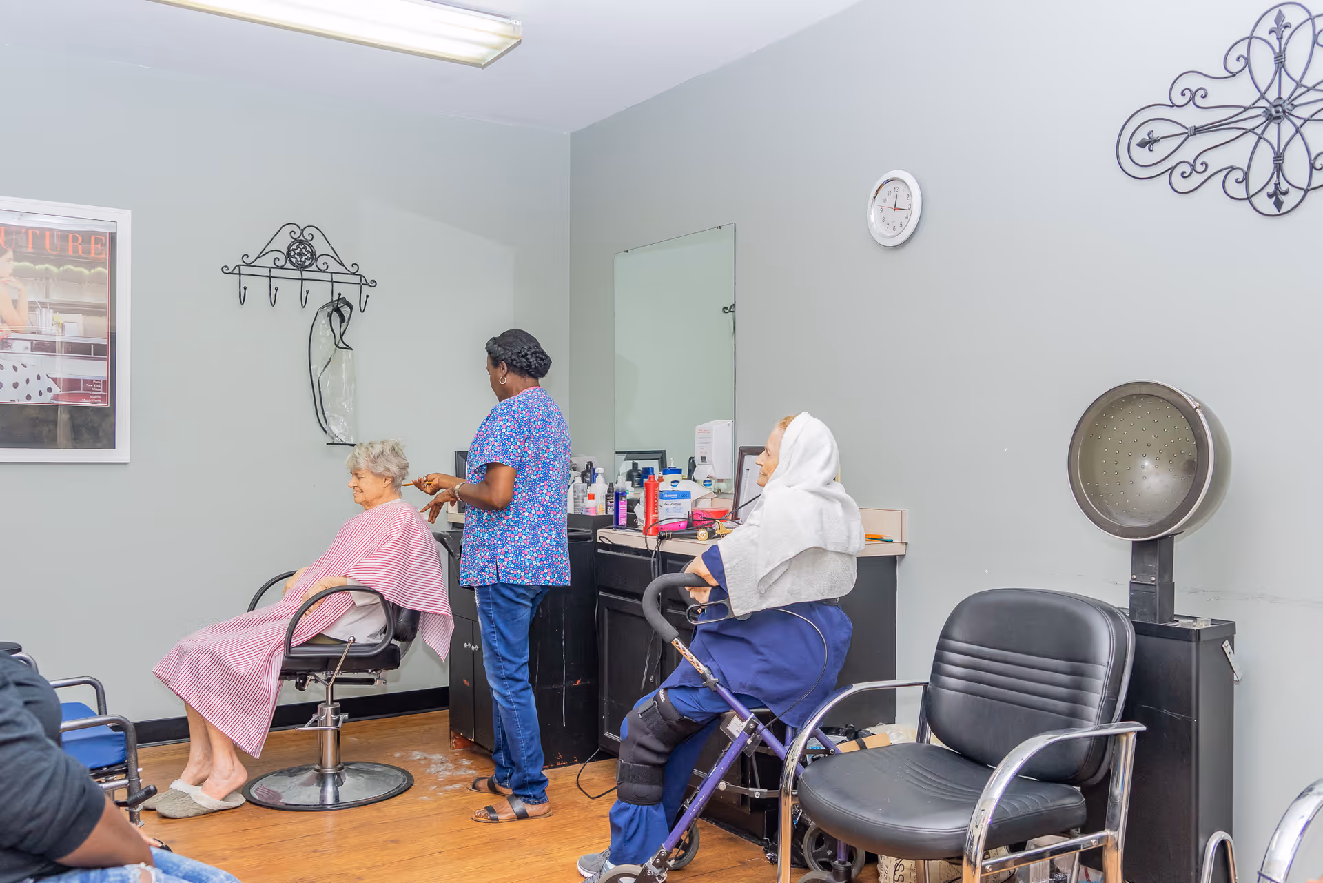 A hair salon inside a nursing and rehabilitation facility where a stylist is cutting an elderly woman's hair. Another elderly woman is seated nearby with a towel over her head, and salon equipment such as chairs, a hair dryer, and hair products are visible.