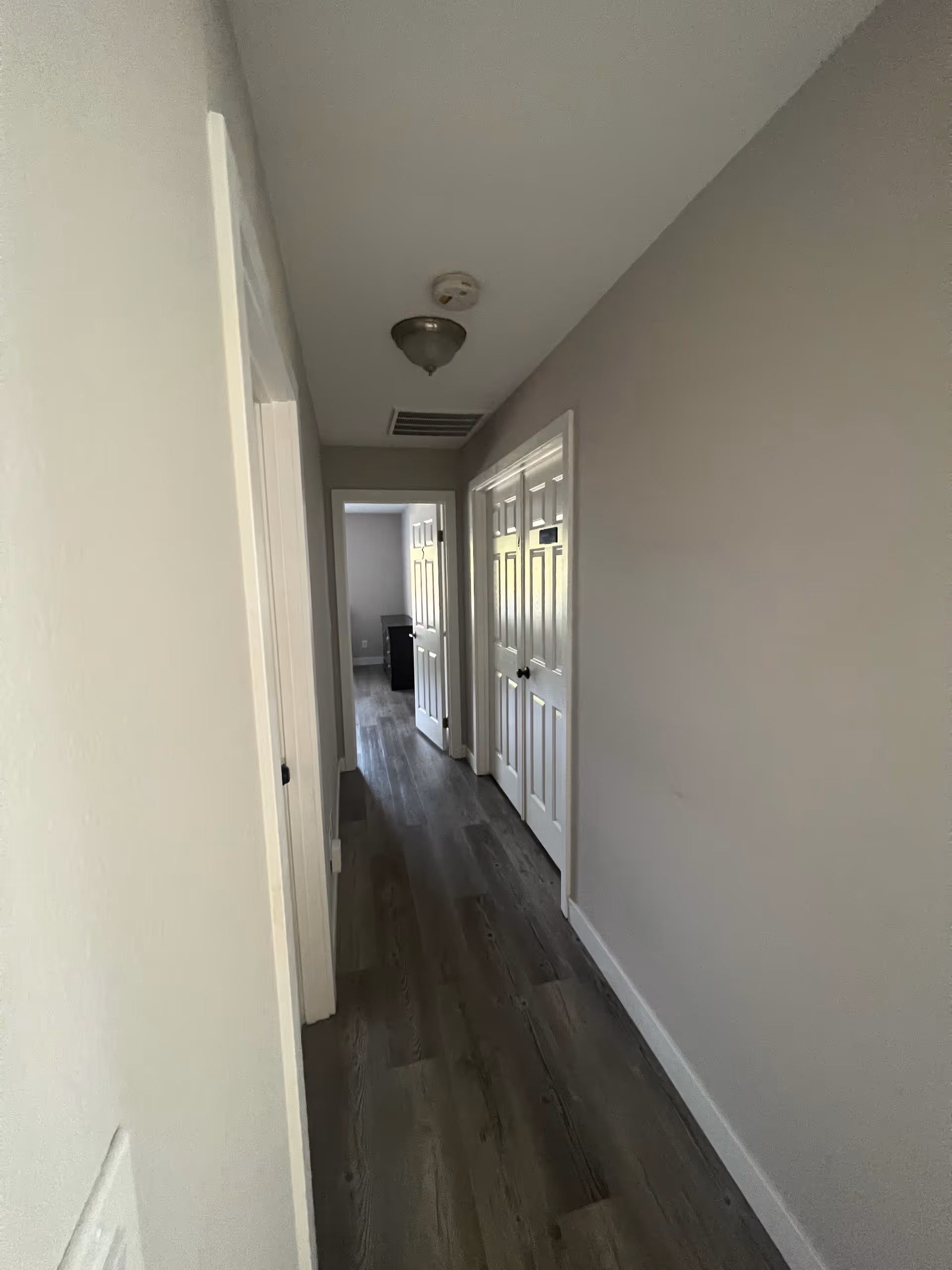 A narrow hallway with light gray walls and dark wood flooring. There are white doors on both sides of the hallway, and a ceiling light fixture is visible. At the end of the hallway, there is an open door leading to another room with a piece of furniture visible.