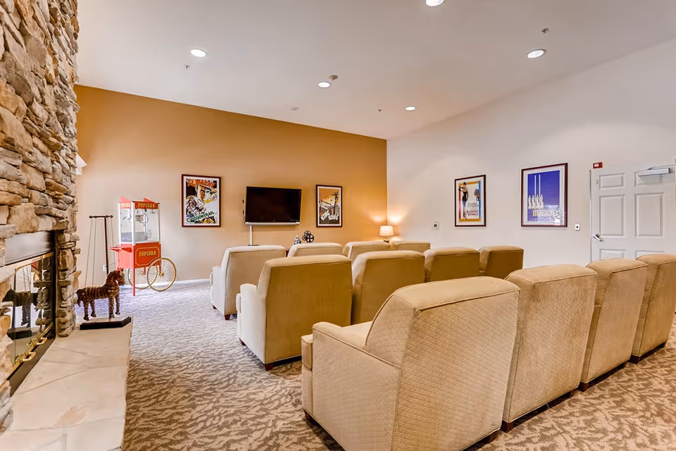 A cozy media room with beige recliner chairs arranged in rows facing a wall-mounted TV, a popcorn cart, stone fireplace, and framed posters.