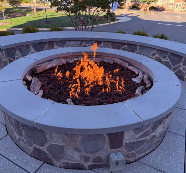 A circular stone fire pit with a burning flame in the center, surrounded by a paved outdoor area with some greenery and a parking lot in the background.