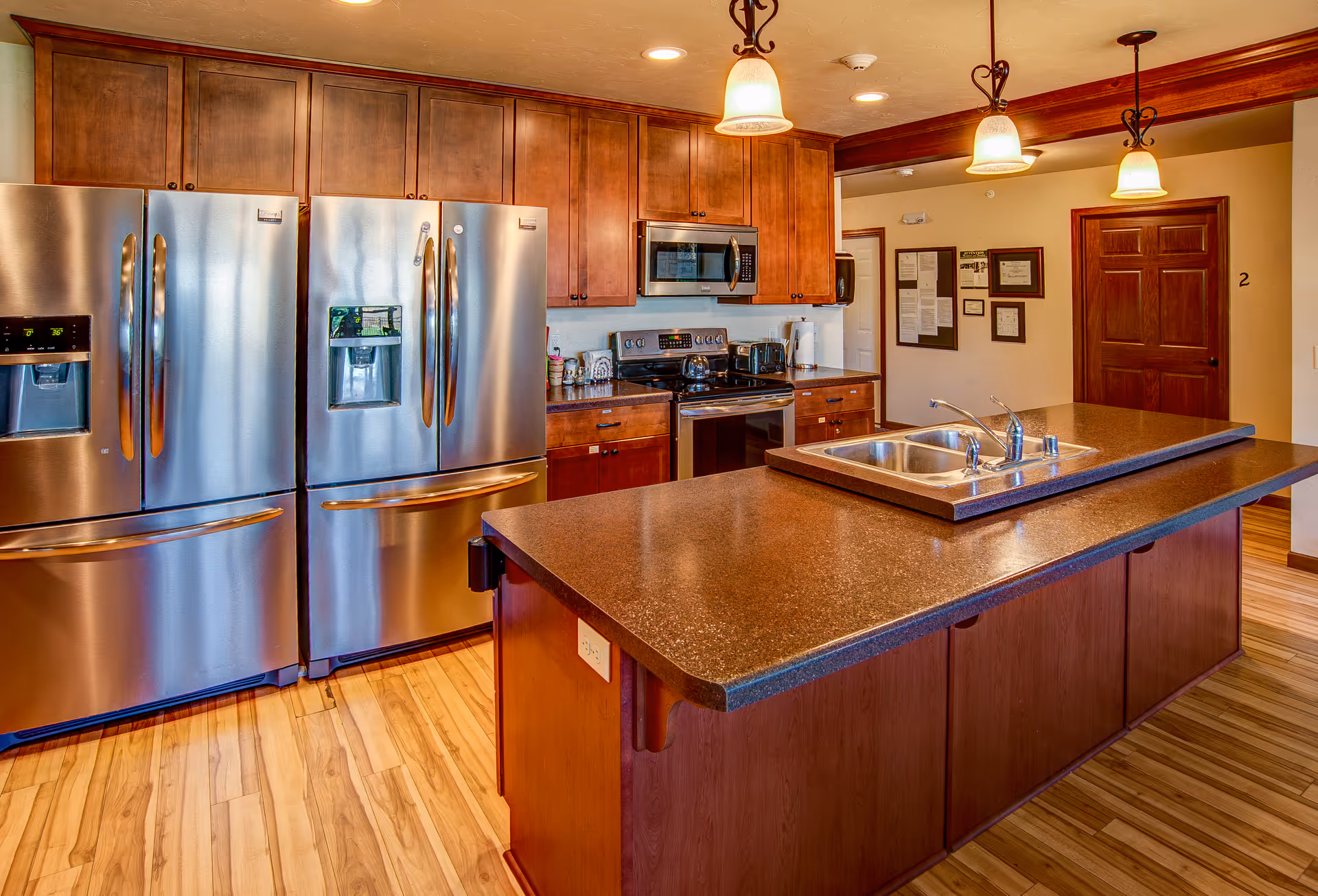 A modern kitchen with wooden cabinets, two stainless steel refrigerators, a stainless steel oven with a microwave above it, and a large kitchen island with a double sink. The floor is wooden, and there are three pendant lights hanging from the ceiling. On the wall in the background, there are framed documents and a wooden door labeled with the number 2.