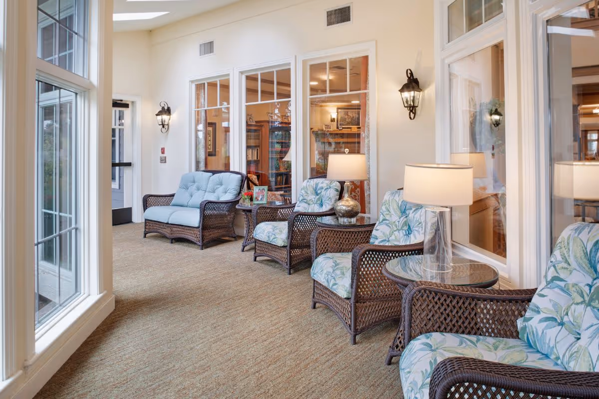 Sunlit lounge with wicker armchairs with floral cushions, side tables and lamps arranged along large windows.