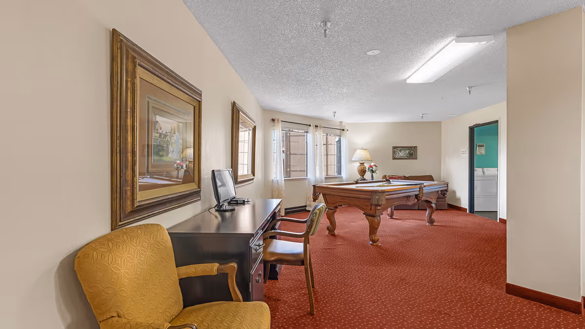 A senior living facility common room with a pool table in the center, a brown couch against the far wall, a table lamp with flowers on a side table, two windows with sheer curtains, a desk with a computer monitor and chair, and a yellow armchair in the foreground. There is also a doorway leading to a laundry room with a washer and dryer visible.