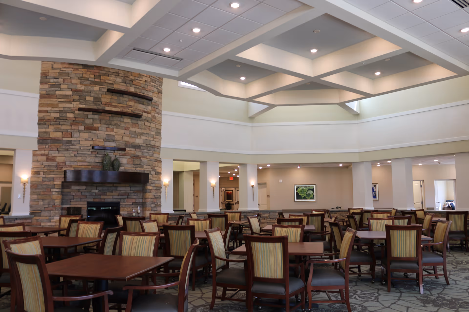 A spacious dining room in a senior living facility with multiple wooden tables and chairs arranged neatly. The room features a high ceiling with recessed lighting and a large stone fireplace with decorative shelves. The walls are painted in neutral tones with framed artwork, and the floor is carpeted with a patterned design.